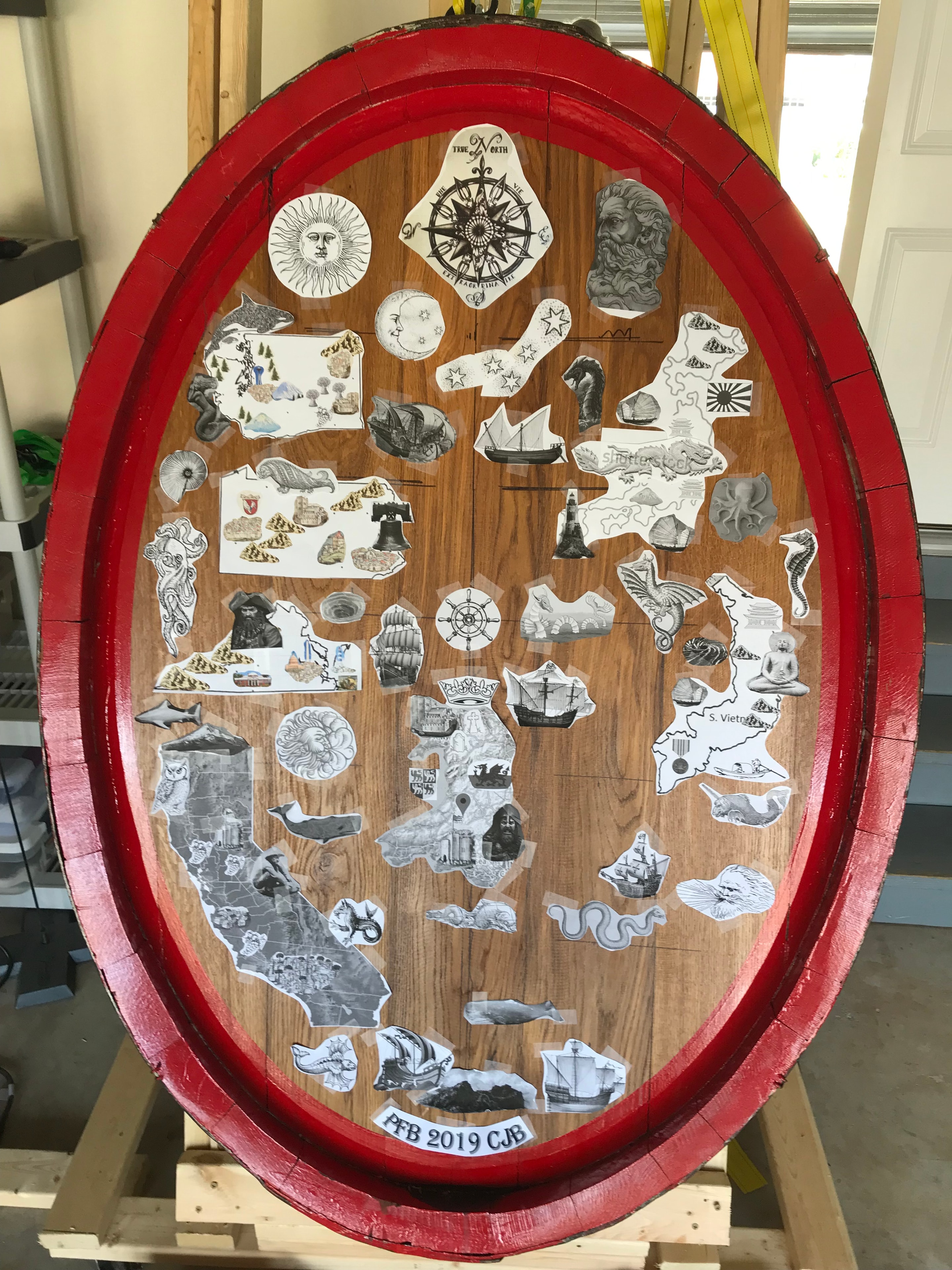 Red-framed oval wine cask end with illustrated design elements laid out on the wood surface