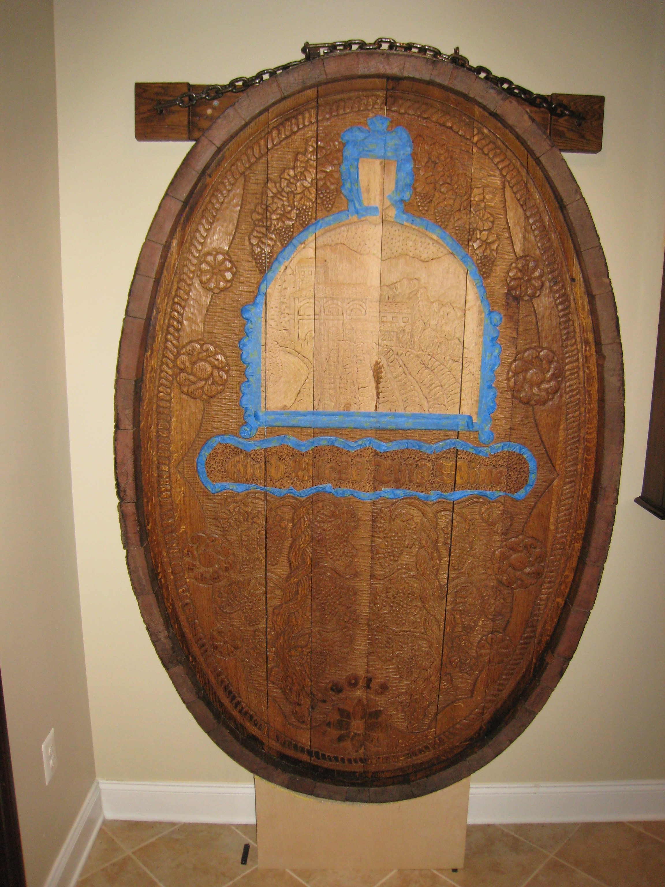The fully carved cask end hanging on the wall in the basement wine area with blue paint markings showing where painting will begin